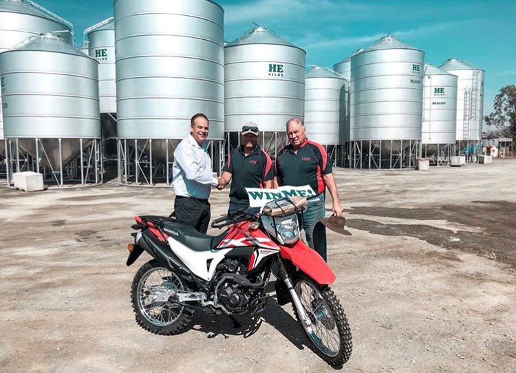 Three men smiling near a motorcycle, holding a "WIN ME!" sign in front of large silos under a clear sky.